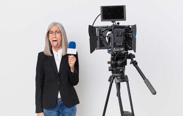 middle age white hair woman with cheerful and rebellious attitude, joking and sticking tongue out and holding a microphone. television presenter concept