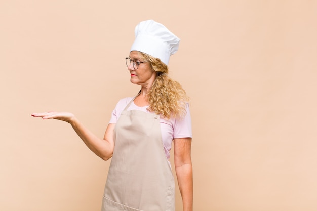 Middle age woman baker feeling happy and smiling casually, looking to an object or concept held on the hand on the side