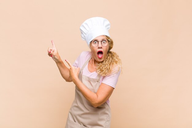 Middle age woman baker feeling joyful and surprised, smiling with a shocked expression and pointing to the side