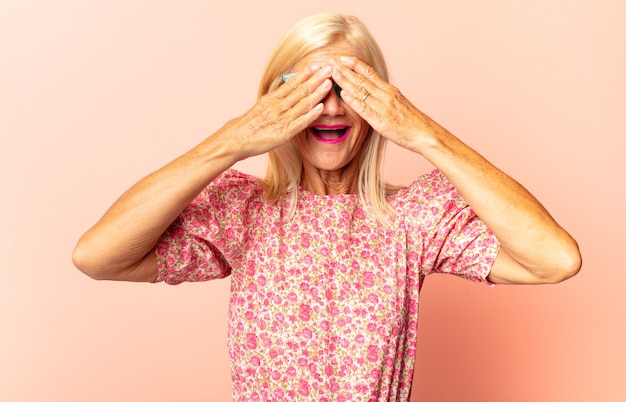 Photo middle age woman covering face with hands, peeking between fingers with surprised expression and looking to the side