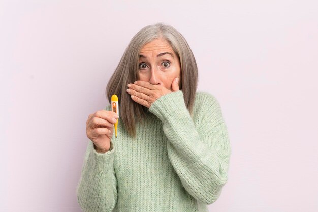 Middle age woman covering mouth with hands with a shocked medicine thermometer concept