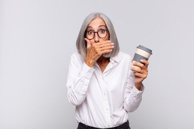 Женщина среднего возраста закрывает рот руками с шокированным, удивленным выражением лица, хранит секрет или говорит о концепции кофе