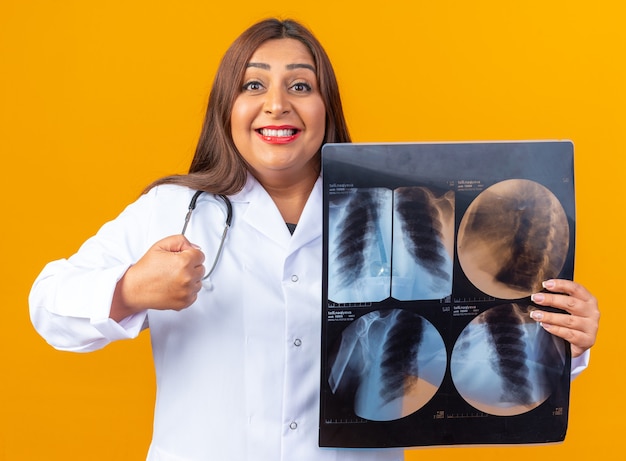 Middle age woman doctor in white coat with stethoscope holding x-ray happy and positive clenching fist