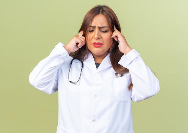 Middle age woman doctor in white coat with stethoscope looking unnoyed touching her temples suffering from headache standing on green