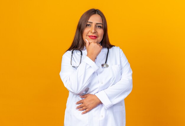 Middle age woman doctor in white coat with stethoscope  with hand on chin smiling confident standing over orange wall