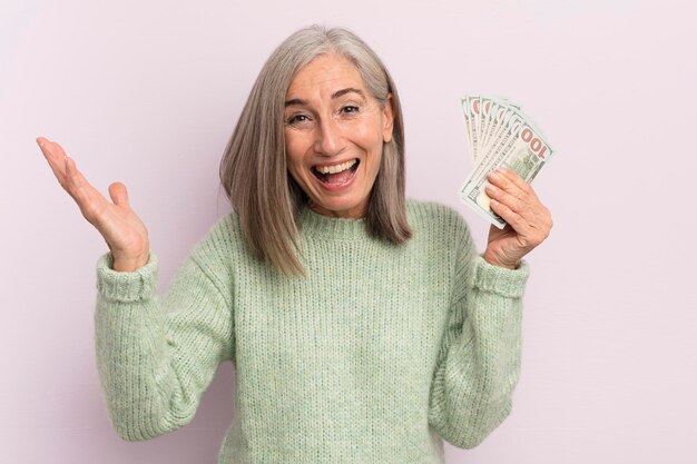 Middle age woman feeling happy and astonished at something unbelievable dollar banknotes concept