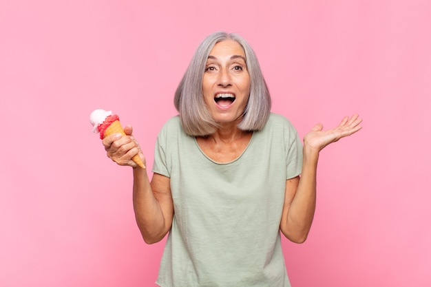 Middle age woman feeling happy, excited, surprised or shocked, smiling and astonished at something unbelievable having an ice cream