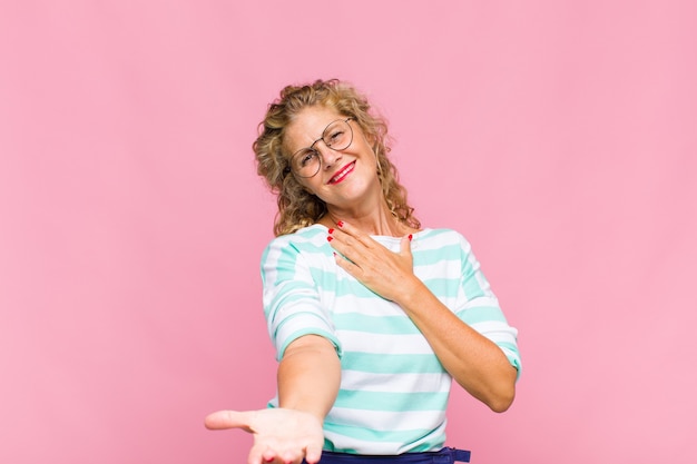 Middle age woman feeling happy and in love, smiling with one hand next to heart and the other stretched up front