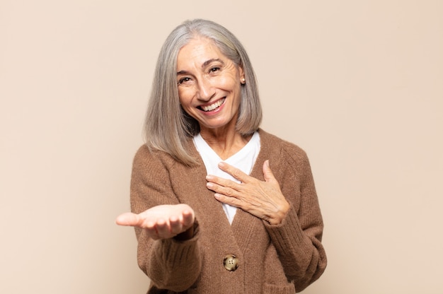 Middle age woman feeling happy and in love, smiling with one hand next to heart and the other stretched up front
