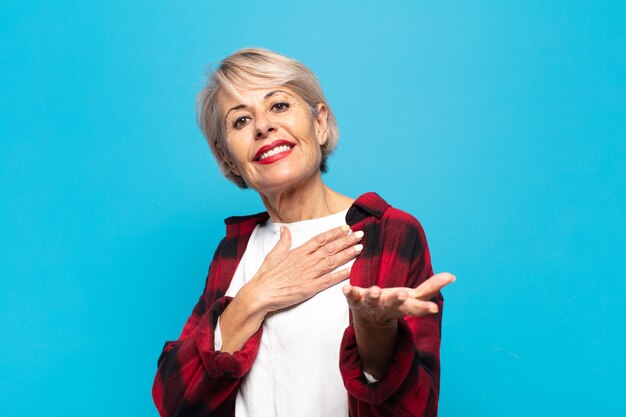 Middle age woman feeling happy and in love, smiling with one hand next to heart and the other stretched up front