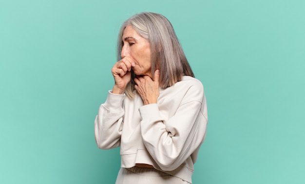 женщина среднего возраста чувствует себя плохо, с болью в горле и симптомами гриппа, кашляет с прикрытым ртом