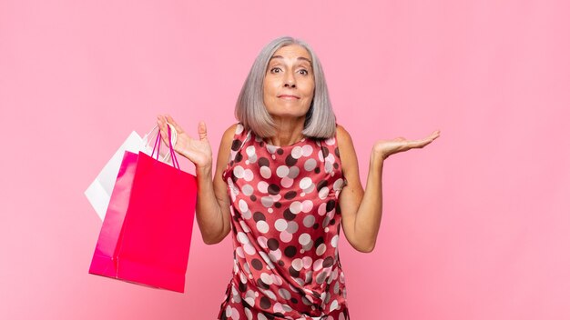 Middle age woman feeling puzzled and confused, doubting, weighting or choosing different options with funny expression with shopping bags