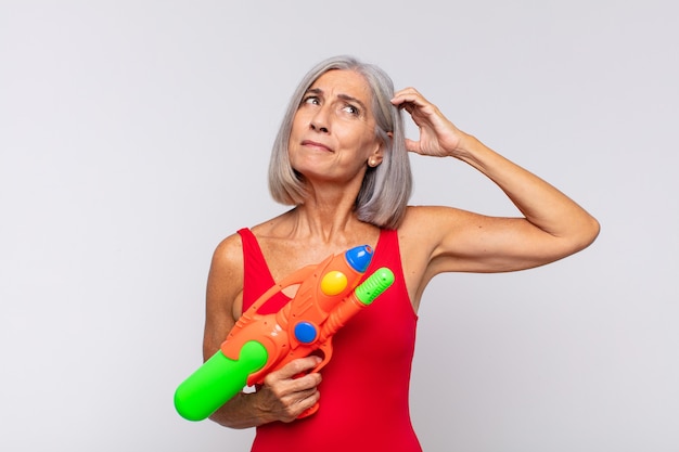 Middle age woman feeling puzzled and confused, scratching head and looking to the side with a water gun