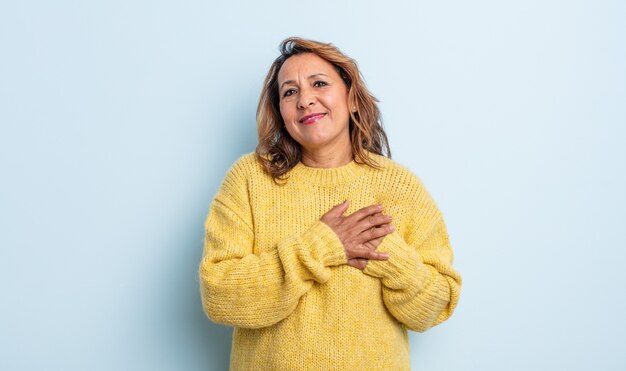 Middle age woman feeling romantic, happy and in love, smiling cheerfully and holding hands close to heart