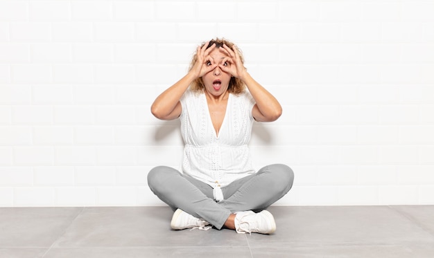 Middle age woman feeling shocked, amazed and surprised, holding glasses with astonished, disbelieving look