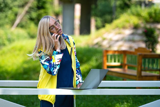 Middle age woman at the garden working from home using laptop and speaking on the phone