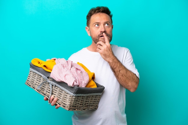 Middle age woman holding a clothes basket isolated on blue background having doubts while looking up