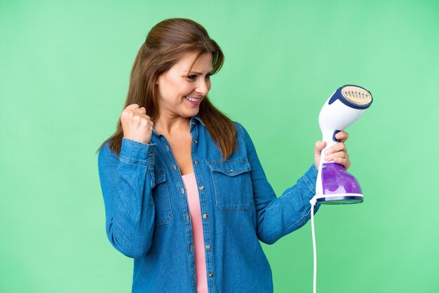 Middle age woman holding an iron over isolated background celebrating a victory