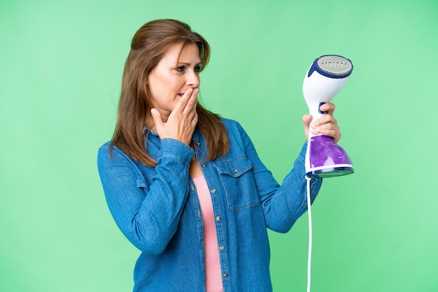 Middle age woman holding an iron over isolated background with surprise and shocked facial expression