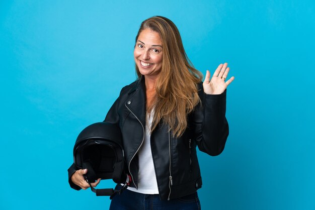 Middle age woman holding a motorcycle helmet isolated on blue saluting with hand with happy expression