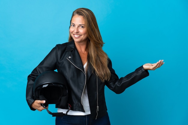 Middle age woman holding a motorcycle helmet isolated on blue wall extending hands to the side for inviting to come
