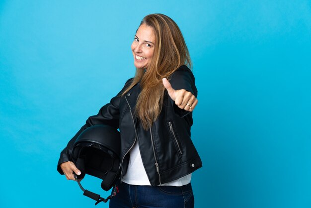 Middle age woman holding a motorcycle helmet isolated on blue wall with thumbs up because something good has happened