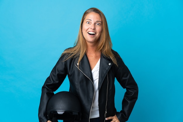 Middle age woman holding a motorcycle helmet isolated on blue with surprise facial expression