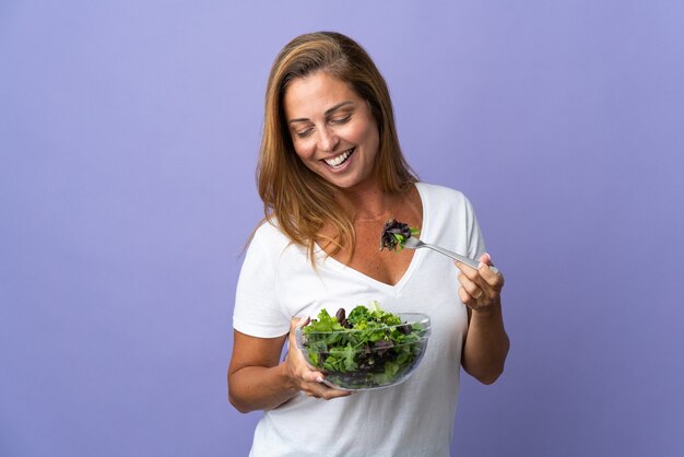 Foto donna di mezza età isolata sulla parete viola che tiene una ciotola di insalata e guardandola con l'espressione felice