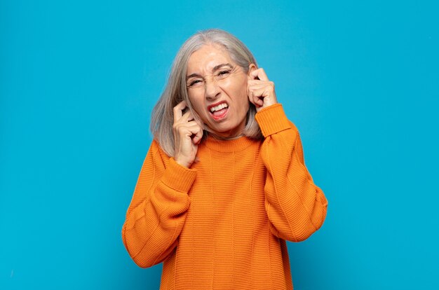 Middle age woman looking angry, stressed and annoyed, covering both ears to a deafening noise, sound or loud music