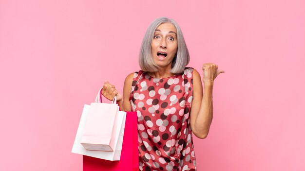 Middle age woman looking astonished in disbelief, pointing at object on the side and saying wow, unbelievable with shopping bags