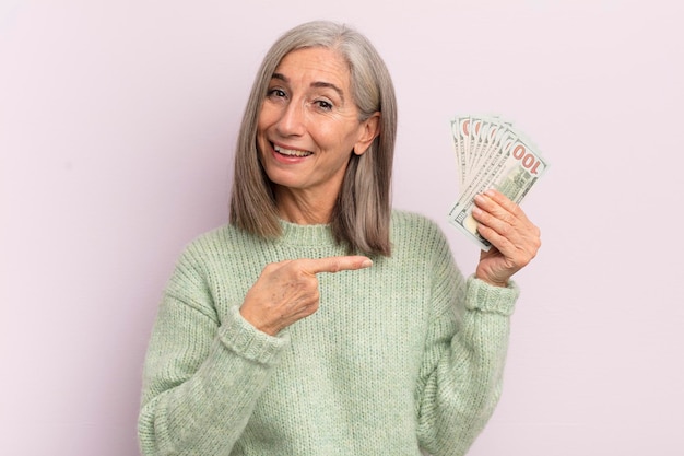 Middle age woman looking excited and surprised pointing to the side dollar banknotes concept