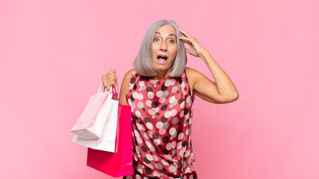 Middle age woman looking happy, astonished and surprised, smiling and realizing amazing and incredible good news with shopping bags