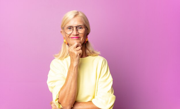 Middle age woman looking happy and smiling with hand on chin, wondering or asking a question, comparing options