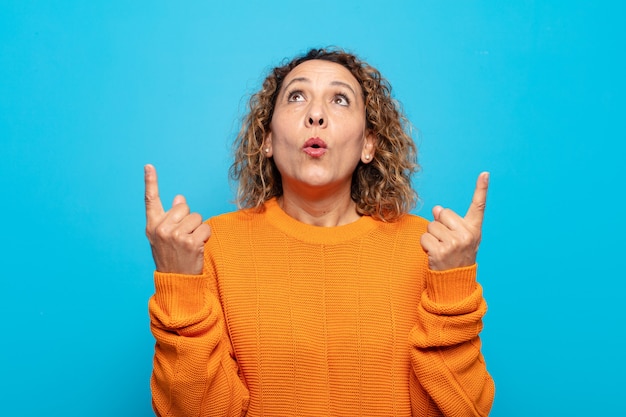 Middle age woman looking shocked, amazed and open mouthed, pointing upwards with both hands to copy space