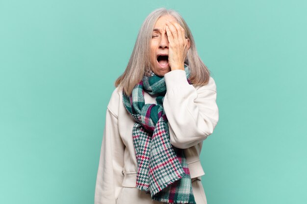 Middle age woman looking sleepy, bored and yawning, with a headache and one hand covering half the face