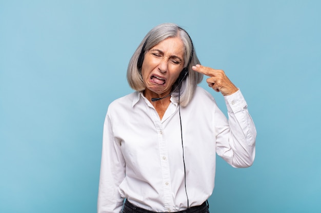 Middle age woman looking unhappy and stressed, suicide gesture making gun sign with hand, pointing to head. telemarketer concept