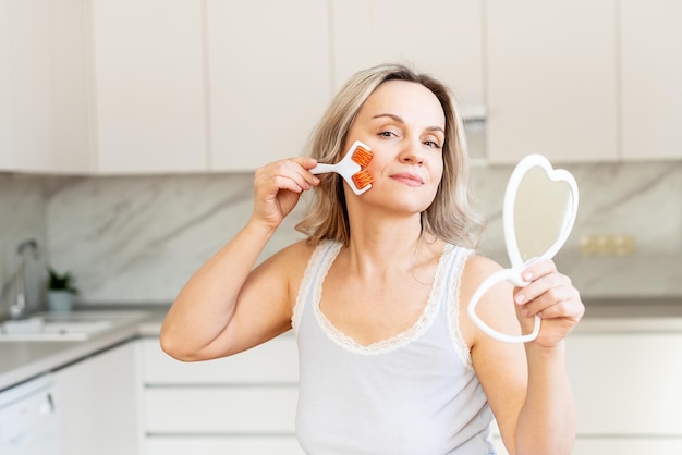 Donna di mezza età in pigiama in cucina con strumento per il massaggio del viso e specchio procedura di bellezza e antietà per la cura della pelle cura del viso cosmetologia della pelle routine mattutina spa e benessere a casa