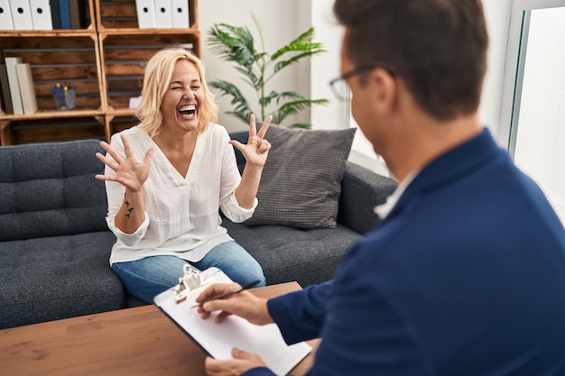 Женщина средних лет в кабинете психологической терапии показывает и указывает пальцами номер восемь, улыбаясь уверенно и счастливо.