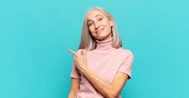 Middle age woman smiling cheerfully, feeling happy and pointing to the side and upwards, showing object in copy space