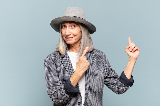 Middle age woman smiling happily and pointing to side and upwards with both hands showing object in copy space