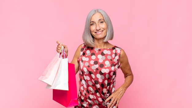 Middle age woman smiling happily with a hand on hip and confident, positive, proud and friendly attitude with shopping bags