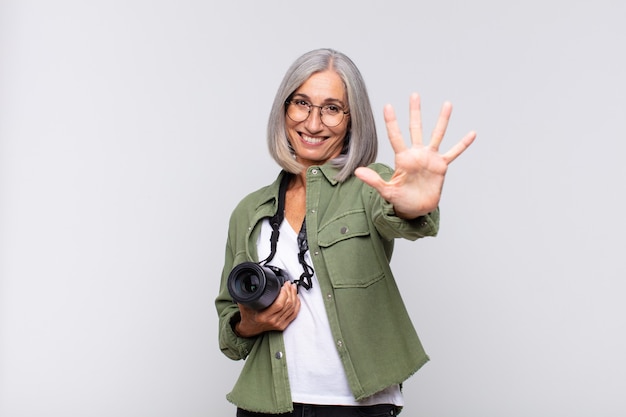 Женщина среднего возраста улыбается и выглядит дружелюбно, показывает номер пять или пятое с рукой вперед, отсчитывая. концепция фотографа