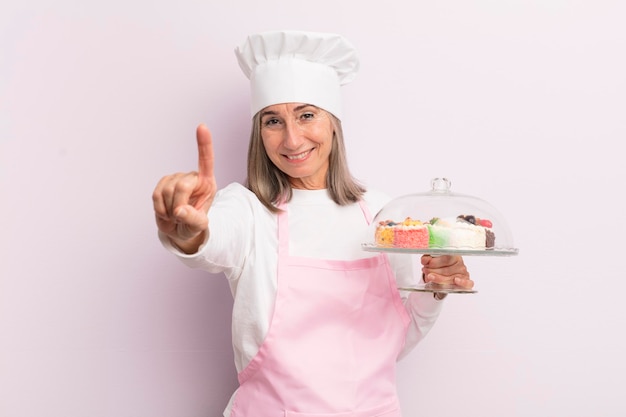 Middle age woman smiling and looking friendly showing number one baker and cakes concept