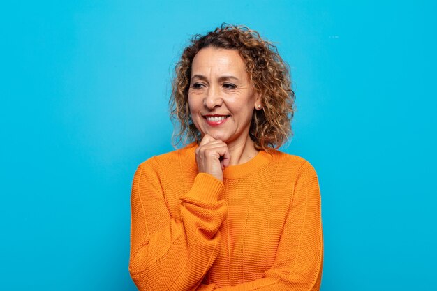 Middle age woman smiling with a happy, confident expression with hand on chin, wondering and looking to the side