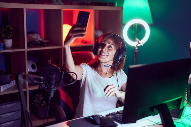 Middle age woman streamer smiling confident make selfie by smartphone at gaming room