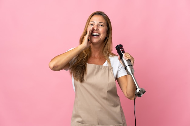 Middle age woman using hand blender isolated on pink shouting with mouth wide open