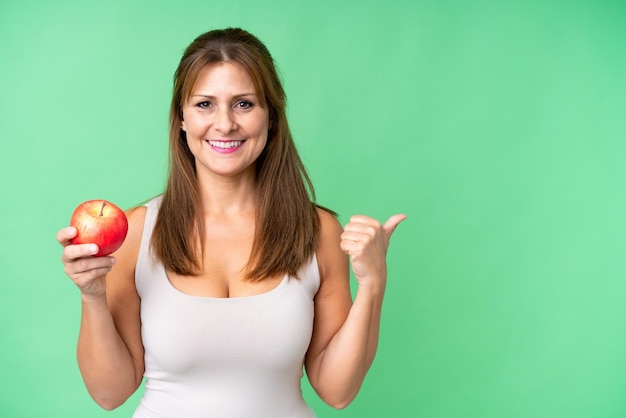 Middle age woman with an apple over isolated background pointing to the side to present a product