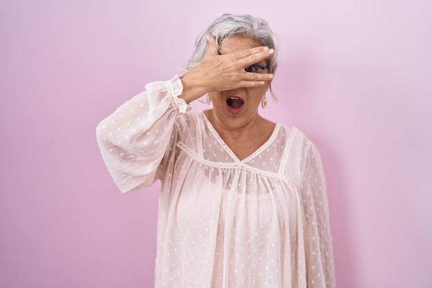 Женщина средних лет с седыми волосами стоит на розовом фоне, заглядывая в шоке, покрывая лицо и глаза рукой, глядя через пальцы с смущенным выражением лица.