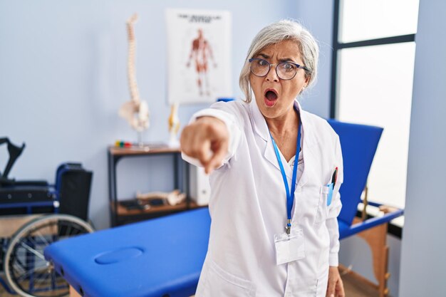 Foto donna di mezza età con i capelli grigi che lavora presso una clinica di recupero dal dolore che punta scontenta e frustrata verso la telecamera arrabbiata e furiosa con te