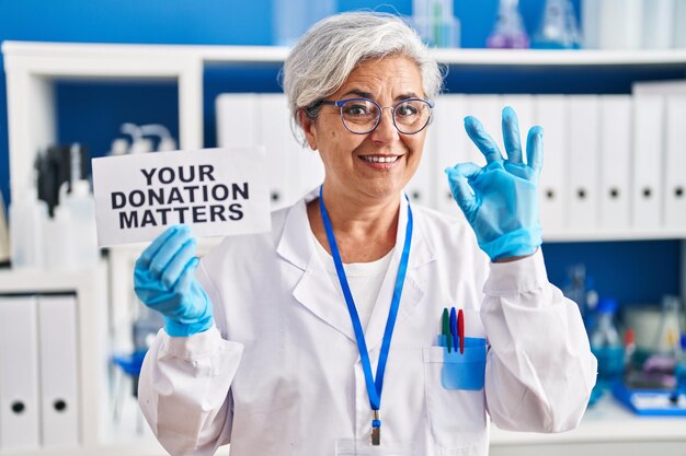 Middle age woman with grey hair working at scientist laboratory holding your donation matters banner doing ok sign with fingers, smiling friendly gesturing excellent symbol